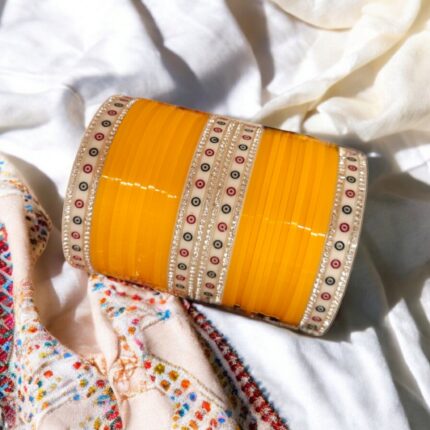 Mango colored Chura with silver gemstones on a white embroidered background.